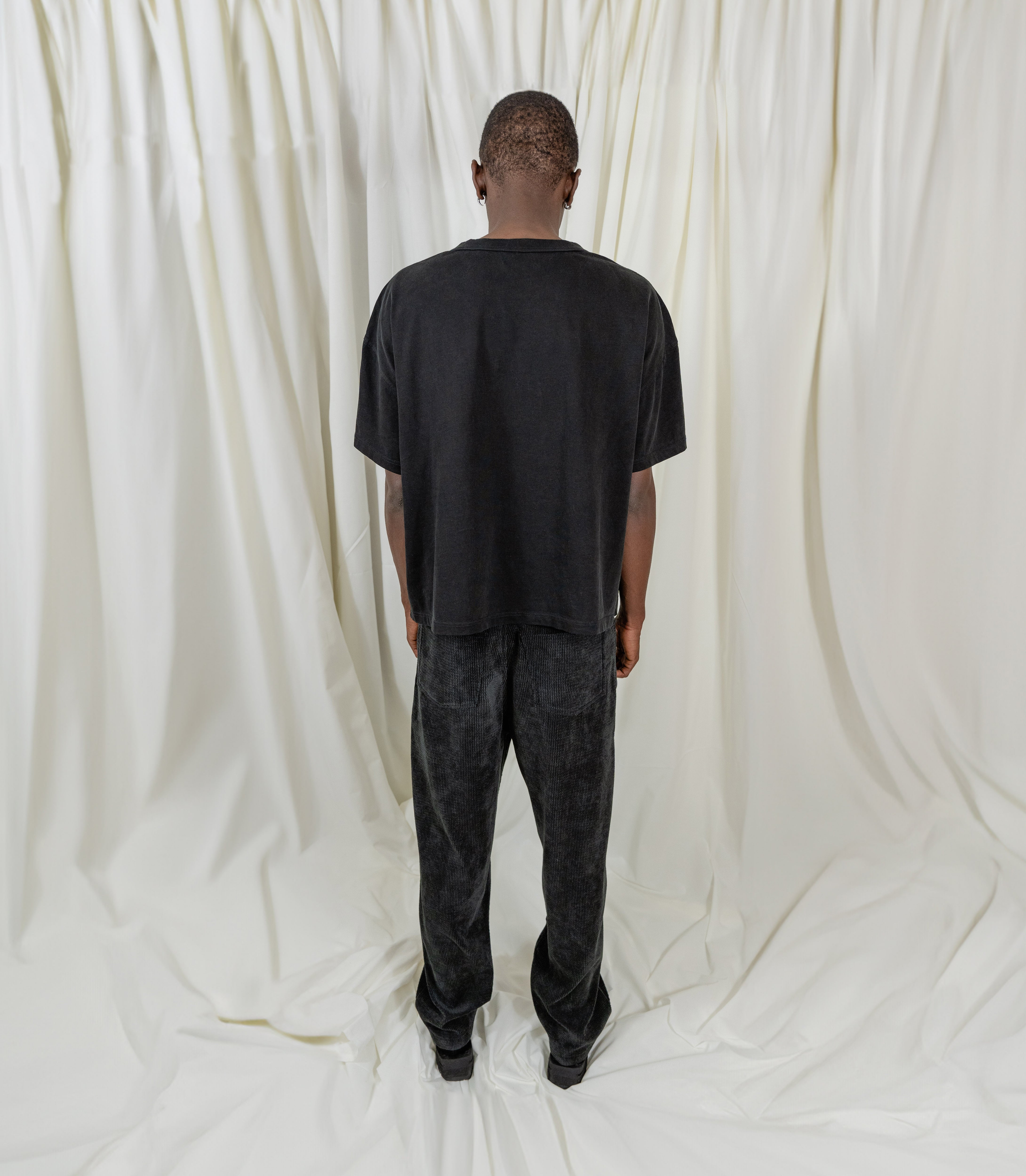 Model Fullsize wearing Signature Peral Tee in Black from Misela Studios in front of a cloth screen - Backview