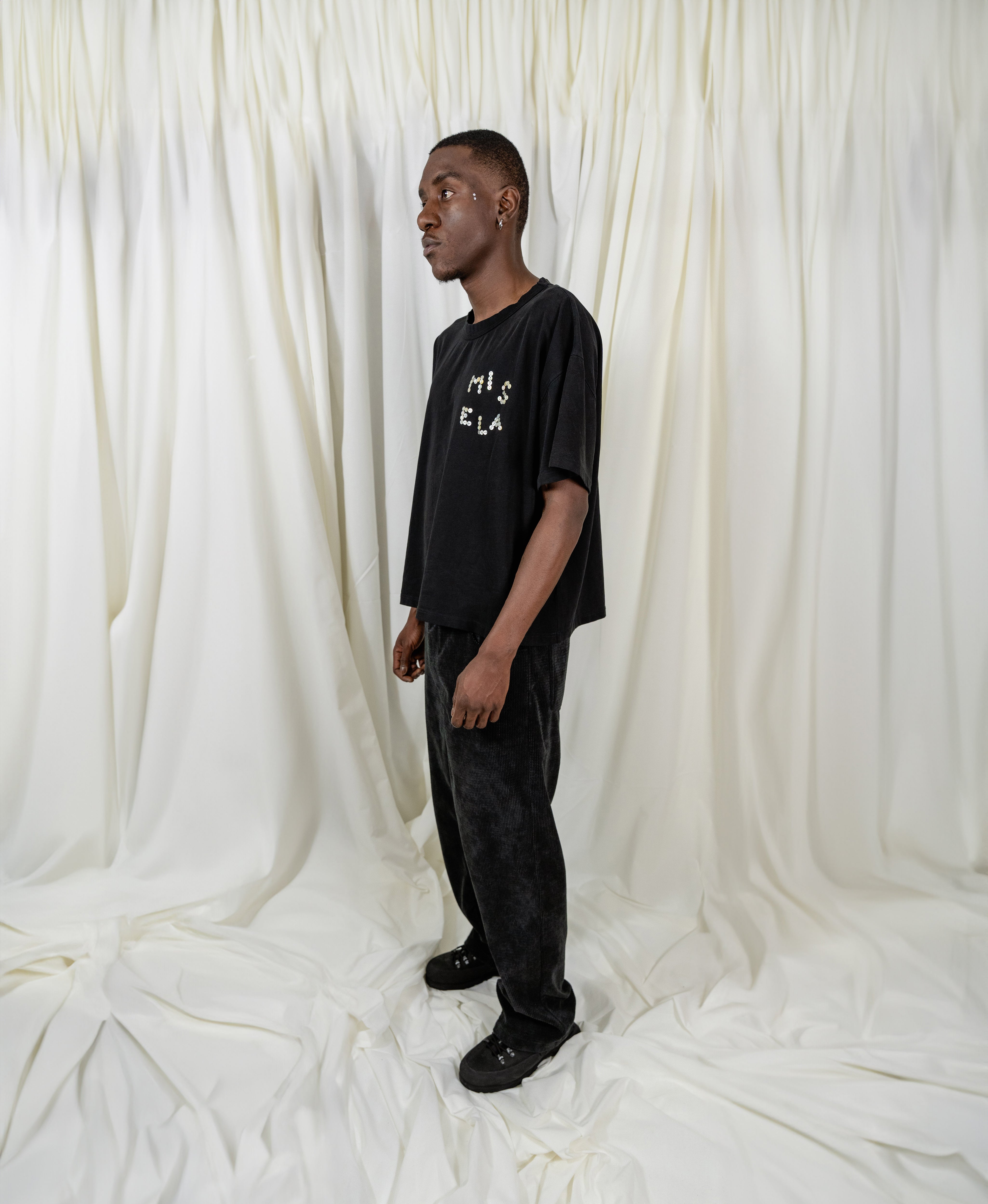 Model Fullsize wearing Signature Peral Tee in Black from Misela Studios in front of a cloth screen - Sideview